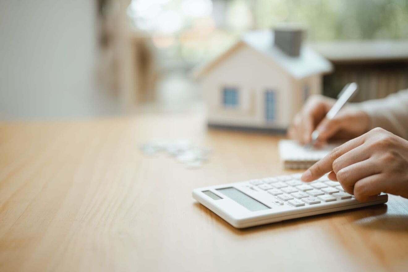 Person using a calculator and taking notes beside a model house, illustrating budgeting before moving to Hamilton.